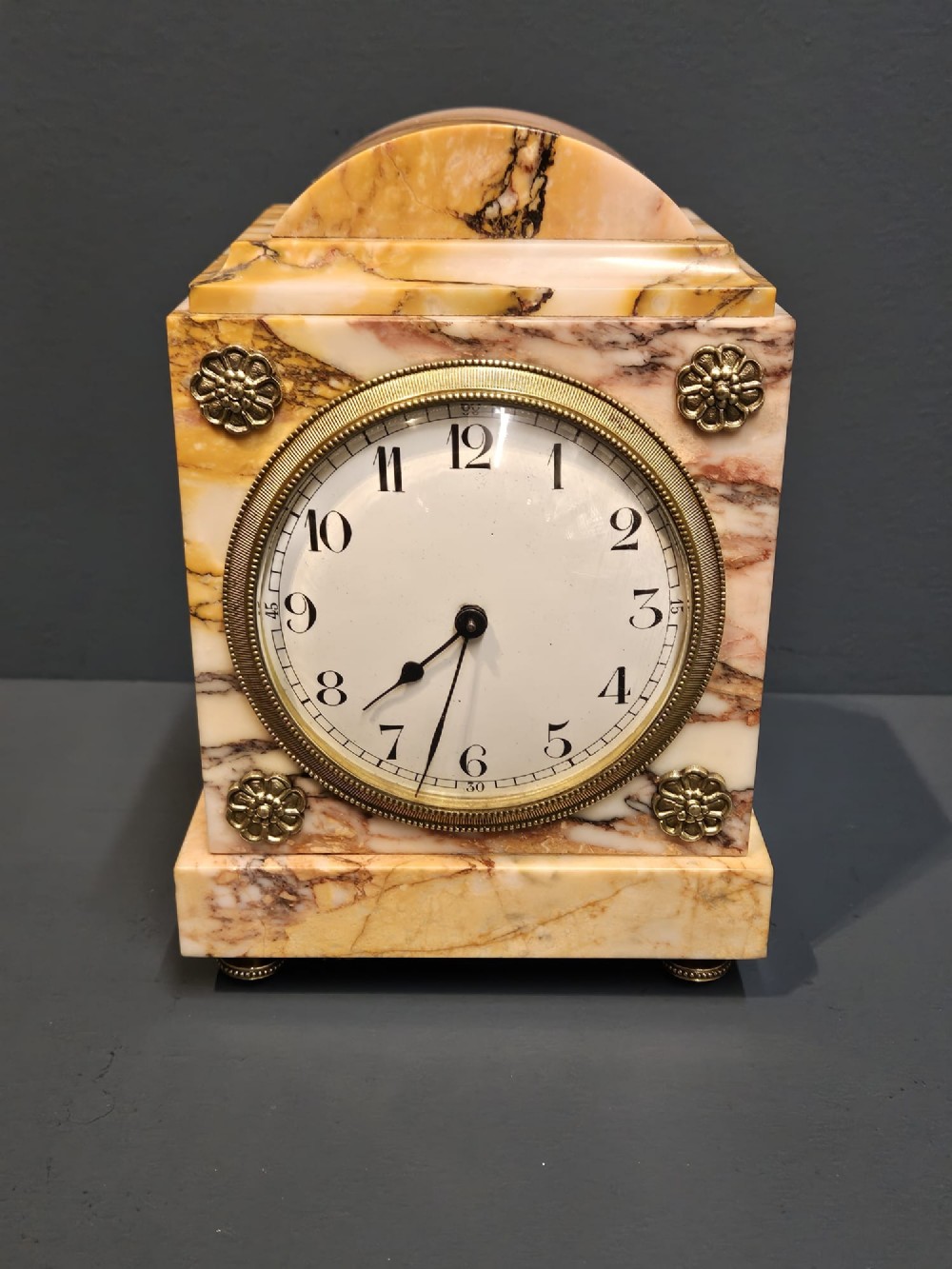 A rectangular Victorian mantel clock crafted from veined cream and brown marble, featuring a domed top, circular brass-rimmed dial with Arabic numerals, and decorative brass corner rosettes.