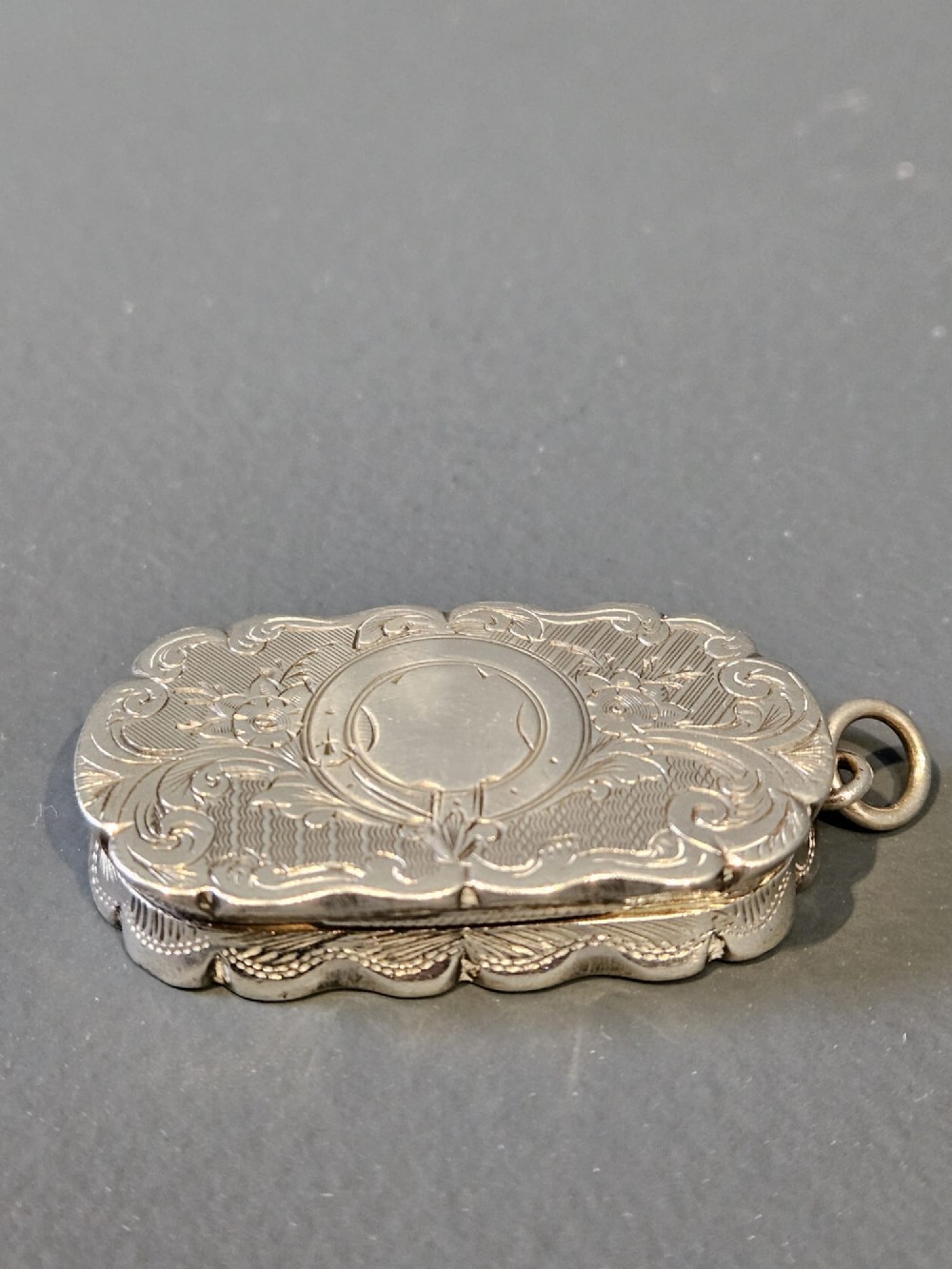 An oblong Victorian silver vinaigrette featuring scalloped edges, intricate floral engraving, an unengraved central cartouche, and an attached suspension loop.