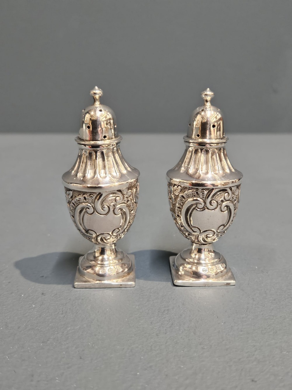 A pair of Victorian sterling silver pepper casters with chased scrollwork decoration and square pedestal bases.