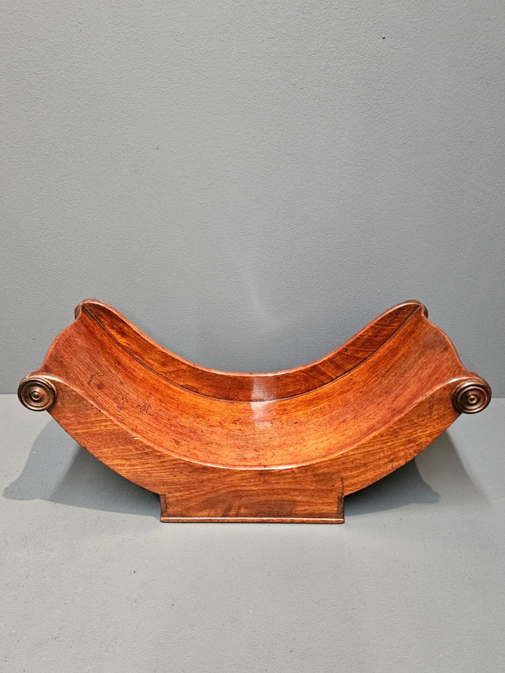 A Regency mahogany cheese coaster featuring a crescent-shaped body with carved scroll terminals on a plinth base.