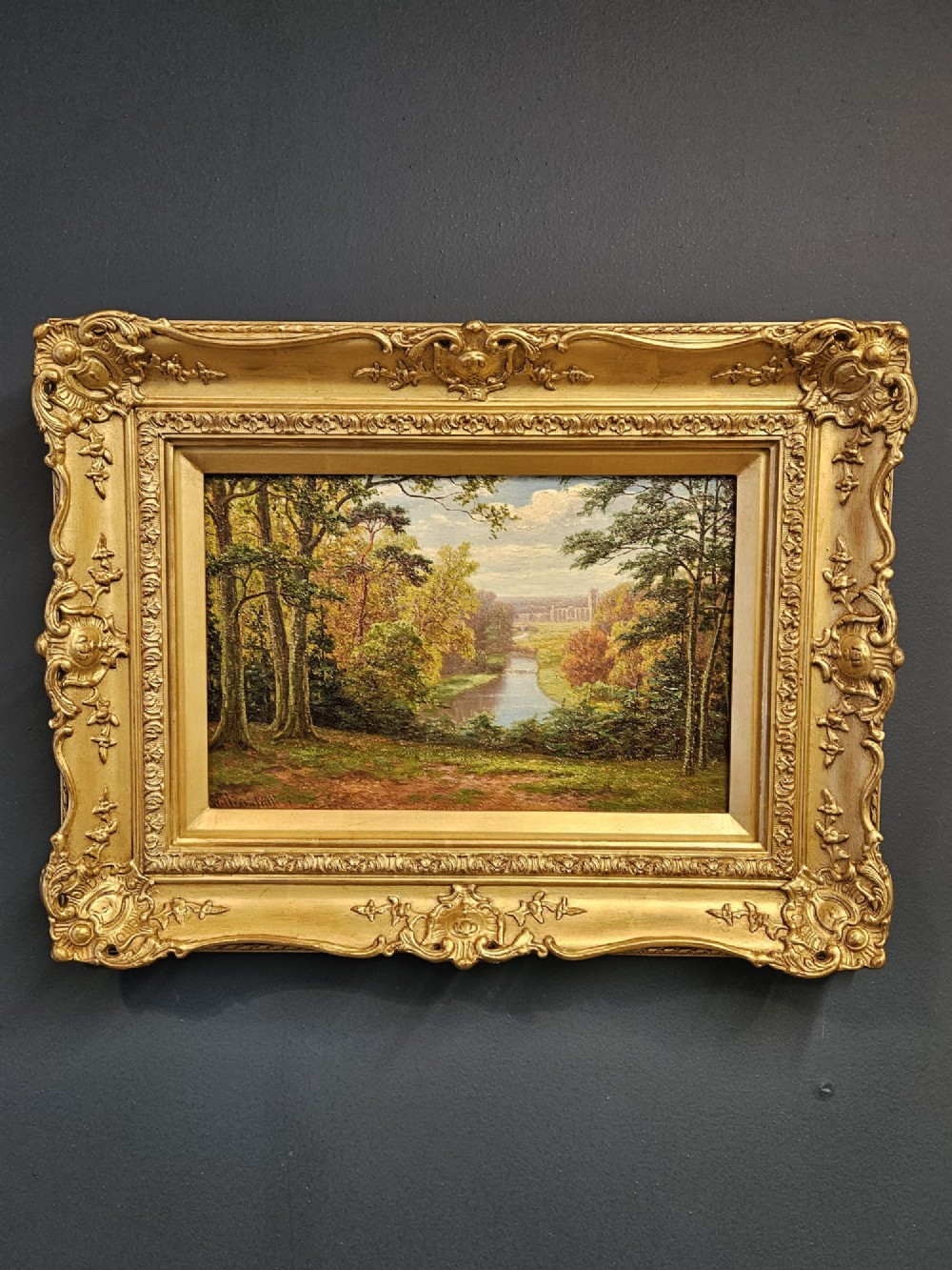 An antique Victorian oil painting depicting a river leading to distant ruins through a sunlit forest, housed in an ornate gilded swept frame.