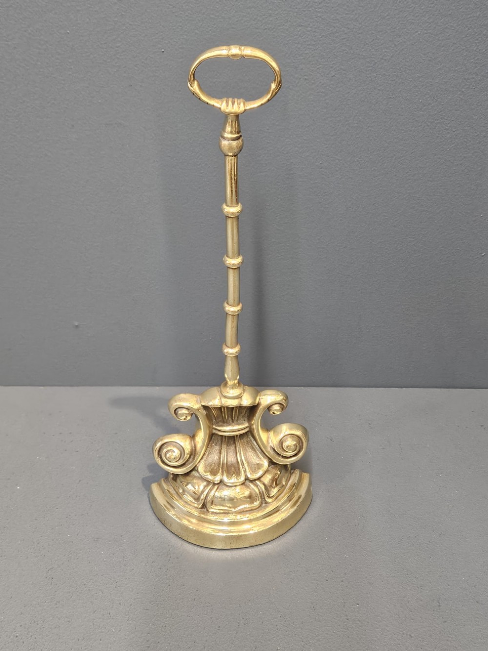A Regency style polished brass door stop featuring a decorative scrolled base, a tall turned stem, and an oval ring handle.