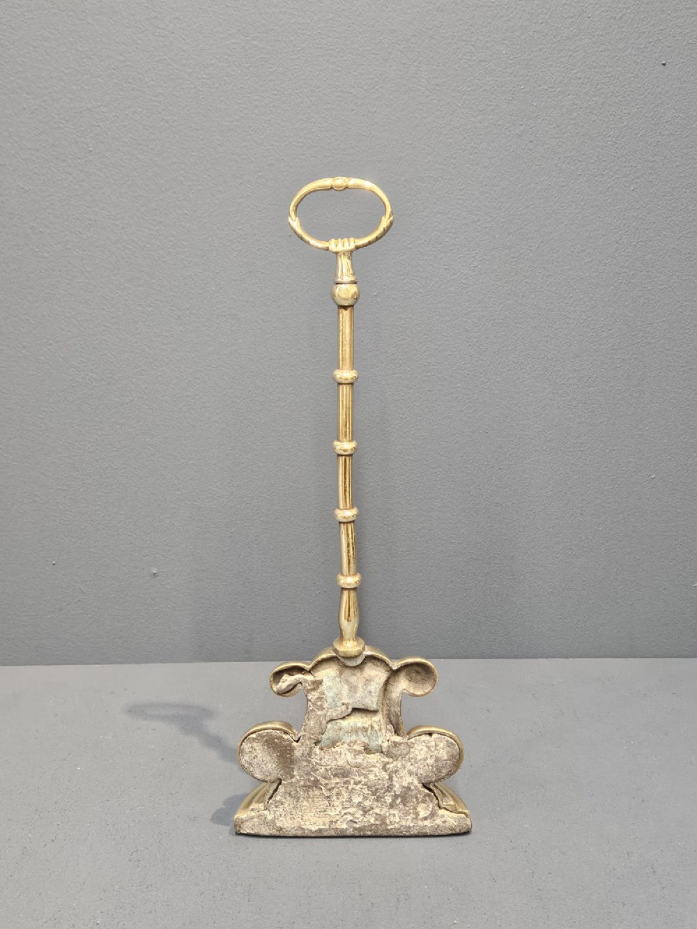 A Regency style polished brass door stop featuring a decorative scrolled base, a tall turned stem, and an oval ring handle. View II