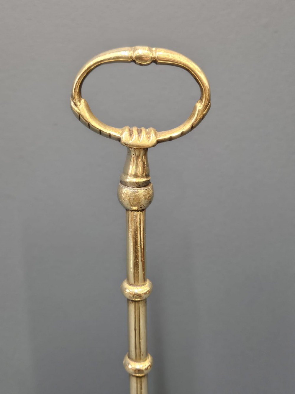 A Regency style polished brass door stop featuring a decorative scrolled base, a tall turned stem, and an oval ring handle. View III