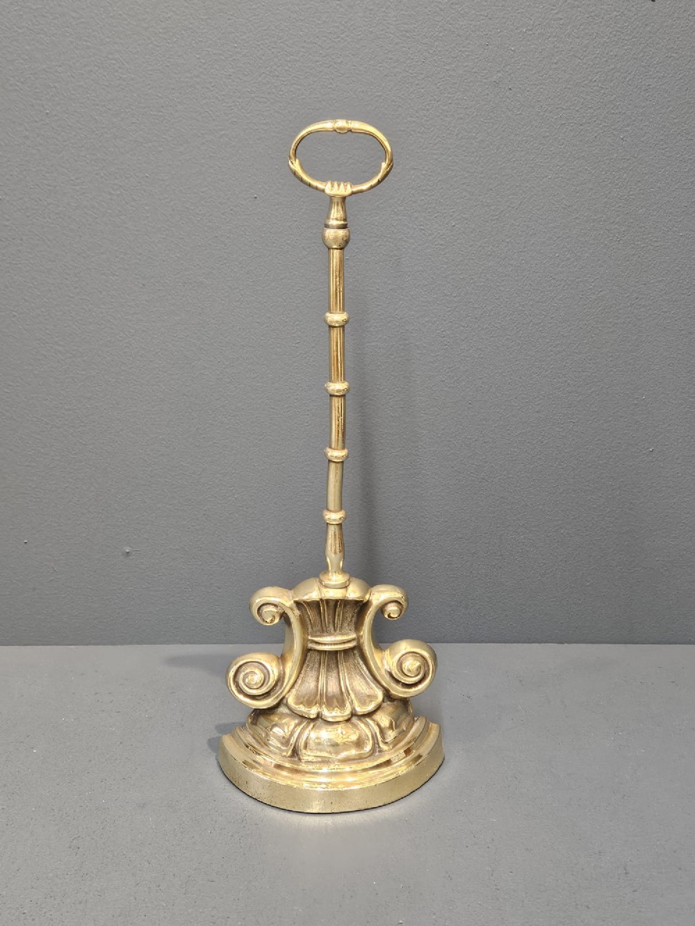 A Regency style polished brass door stop featuring a decorative scrolled base, a tall turned stem, and an oval ring handle. View VI