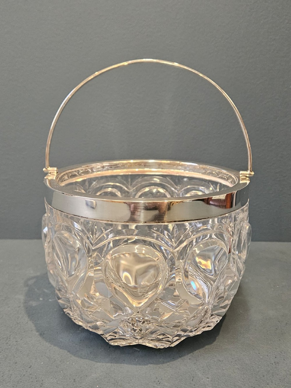 A clear cut crystal ice pail featuring an arched decorative motif, fitted with a silver rim and a slender swing handle.