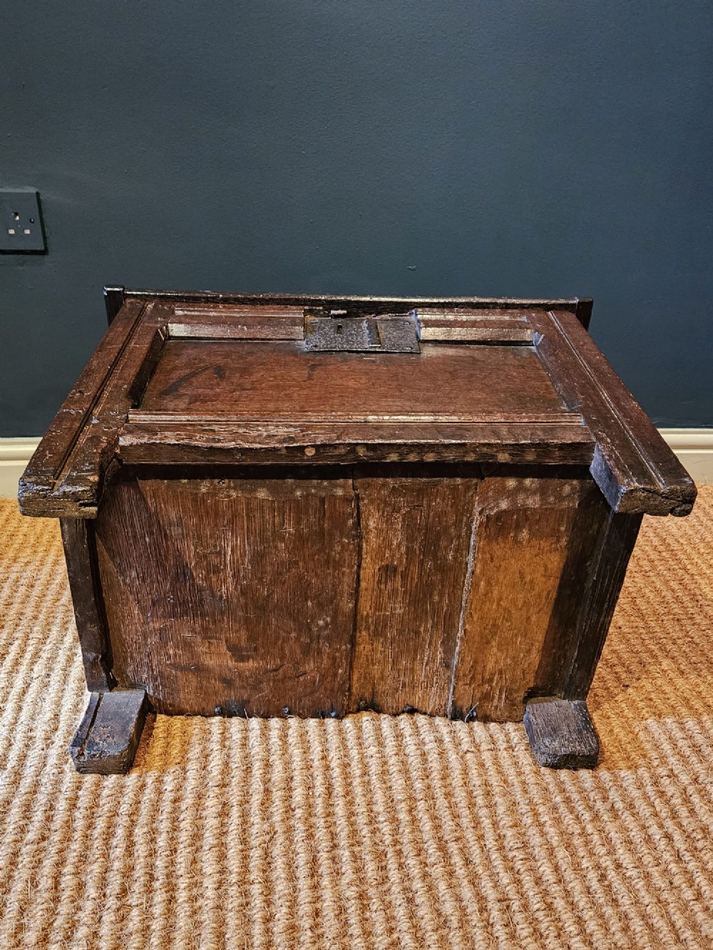 A small 17th-century dark oak coffer featuring a single front panel, a hinged plank top, and a wrought iron lock plate. View II