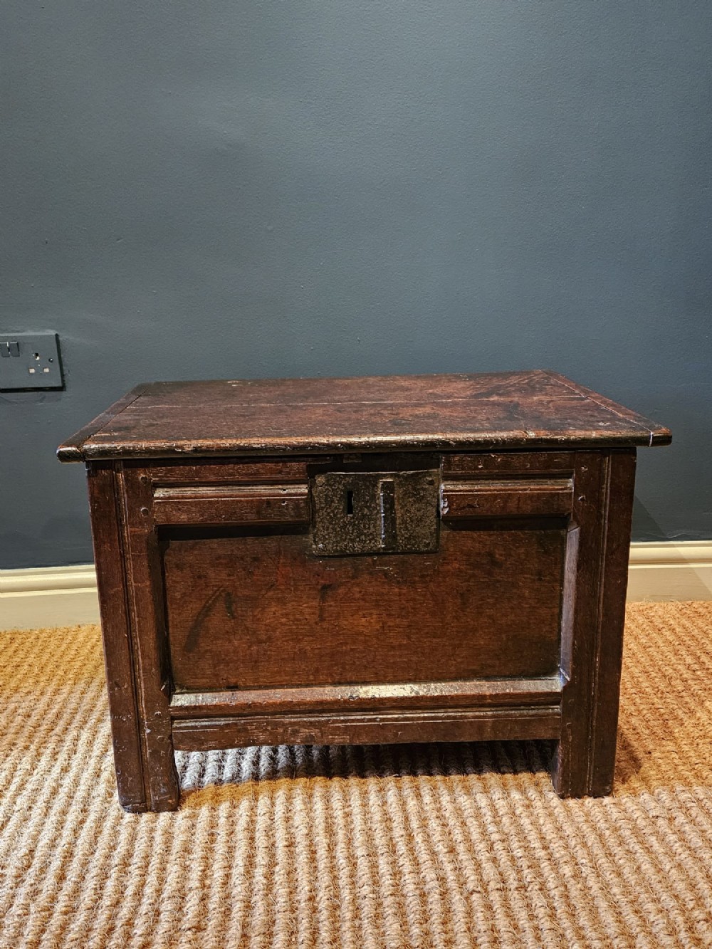 A small 17th-century dark oak coffer featuring a single front panel, a hinged plank top, and a wrought iron lock plate. View III