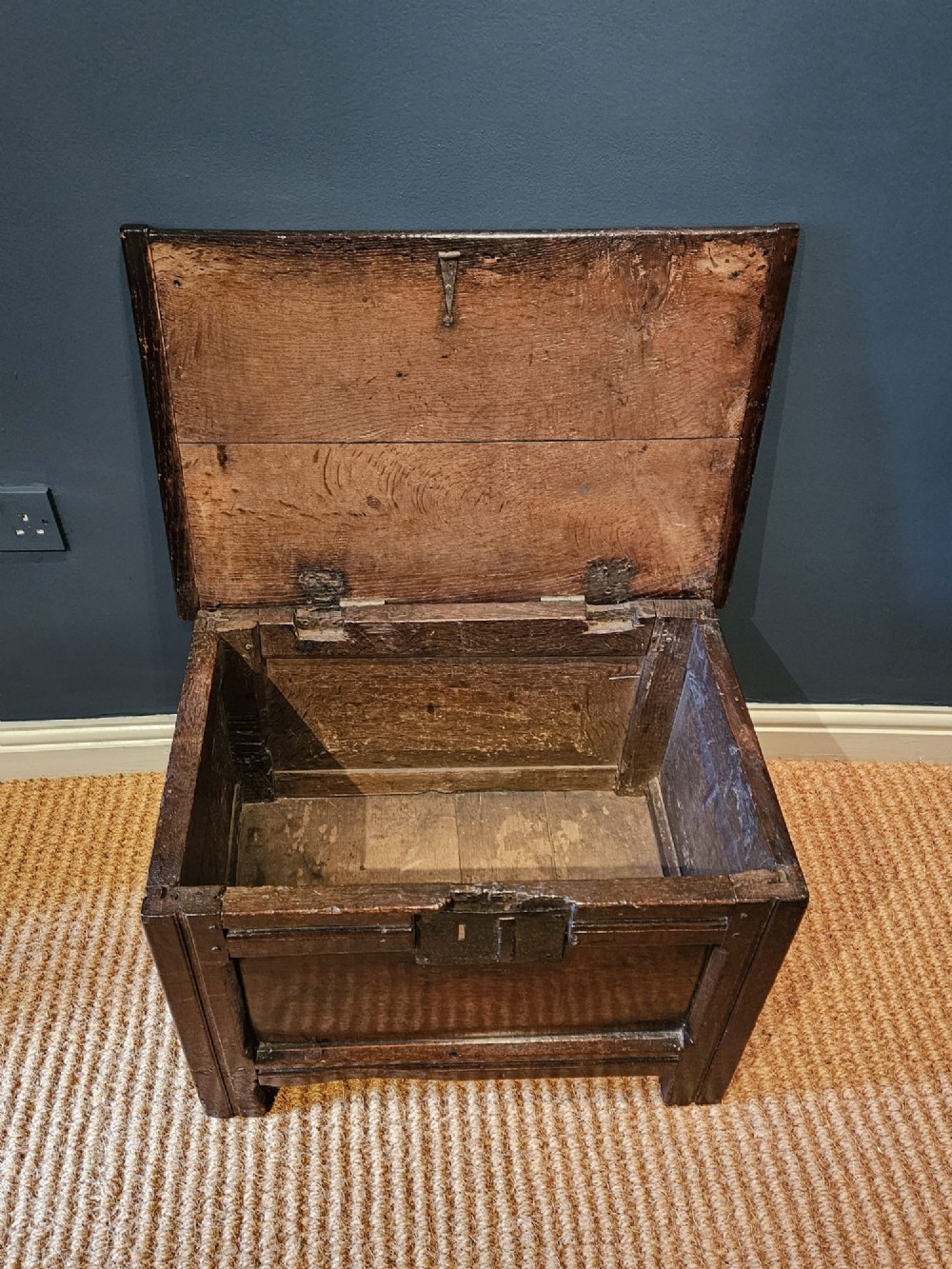 A small 17th-century dark oak coffer featuring a single front panel, a hinged plank top, and a wrought iron lock plate. View V
