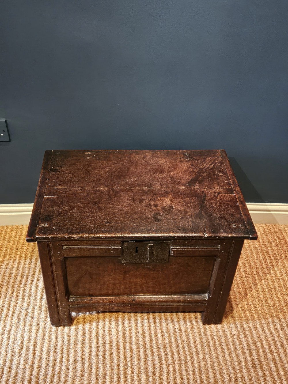 A small 17th-century dark oak coffer featuring a single front panel, a hinged plank top, and a wrought iron lock plate. View VI