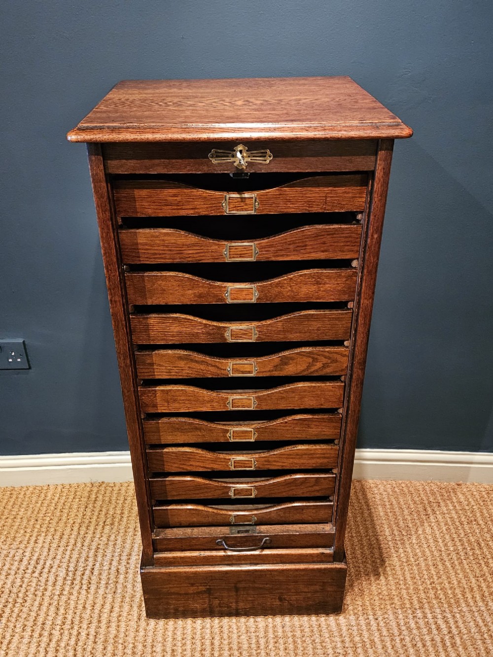 A tall Victorian medium brown oak filing cabinet featuring internal document trays with brass label holders and a top key lock.