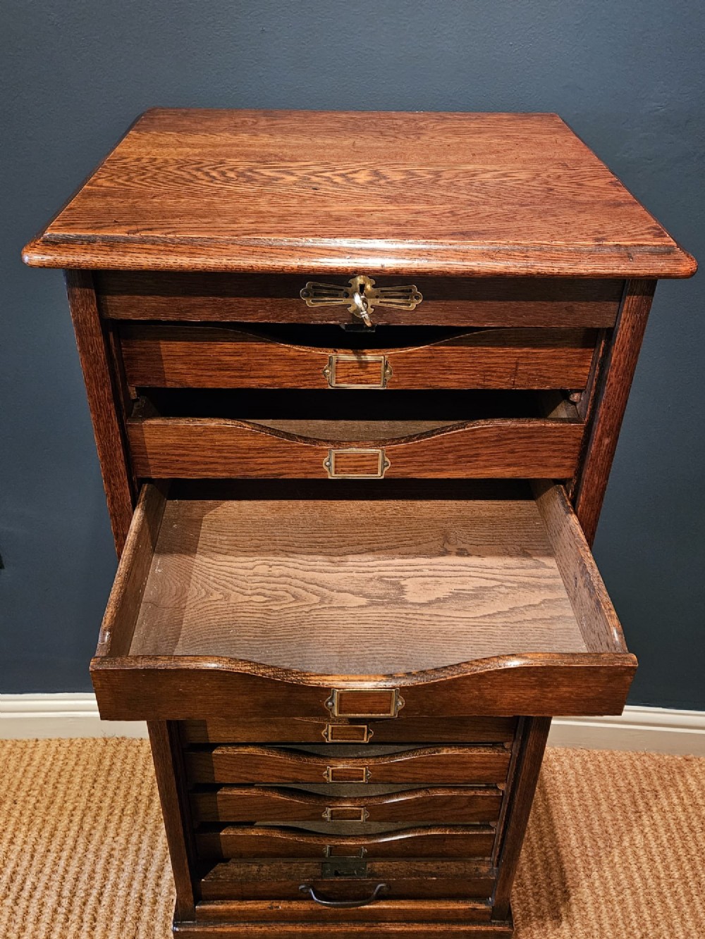 A tall Victorian medium brown oak filing cabinet featuring internal document trays with brass label holders and a top key lock. View IV