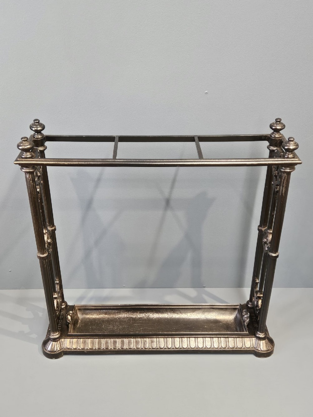 A rectangular Victorian cast iron stick stand featuring corner finials, multi-compartment top rail, vertical column supports, and a drip tray base.