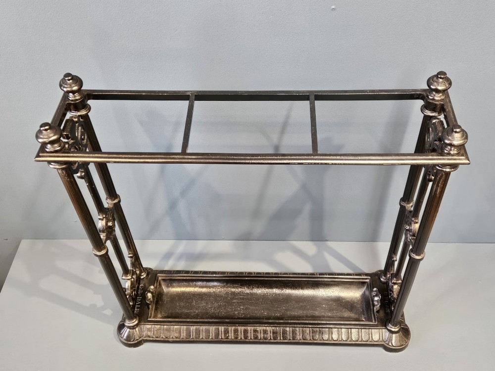 A rectangular Victorian cast iron stick stand featuring corner finials, multi-compartment top rail, vertical column supports, and a drip tray base. View II