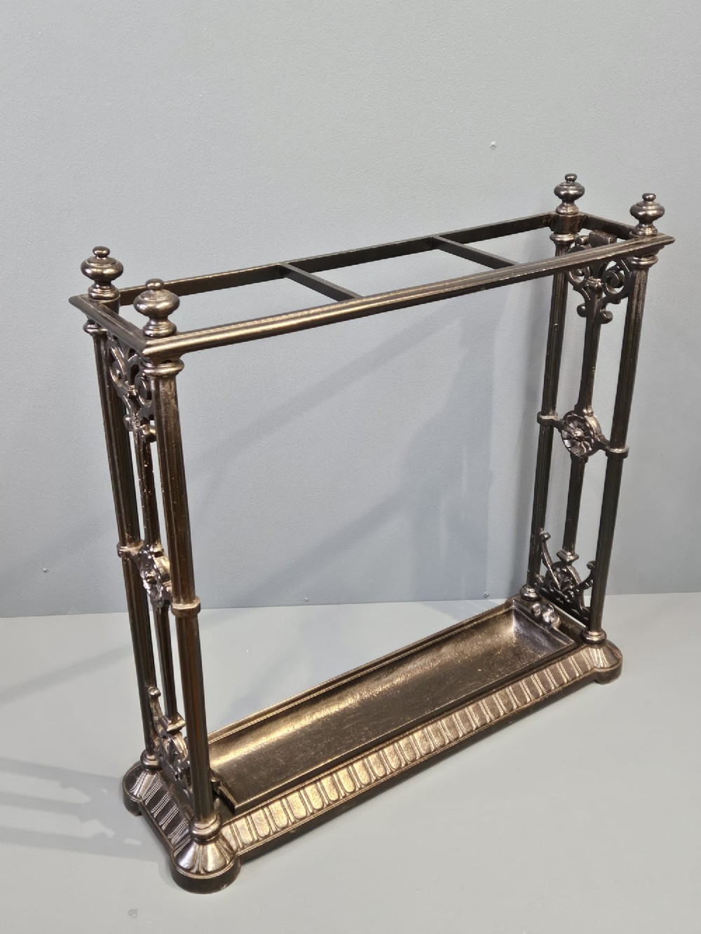 A rectangular Victorian cast iron stick stand featuring corner finials, multi-compartment top rail, vertical column supports, and a drip tray base. View IV