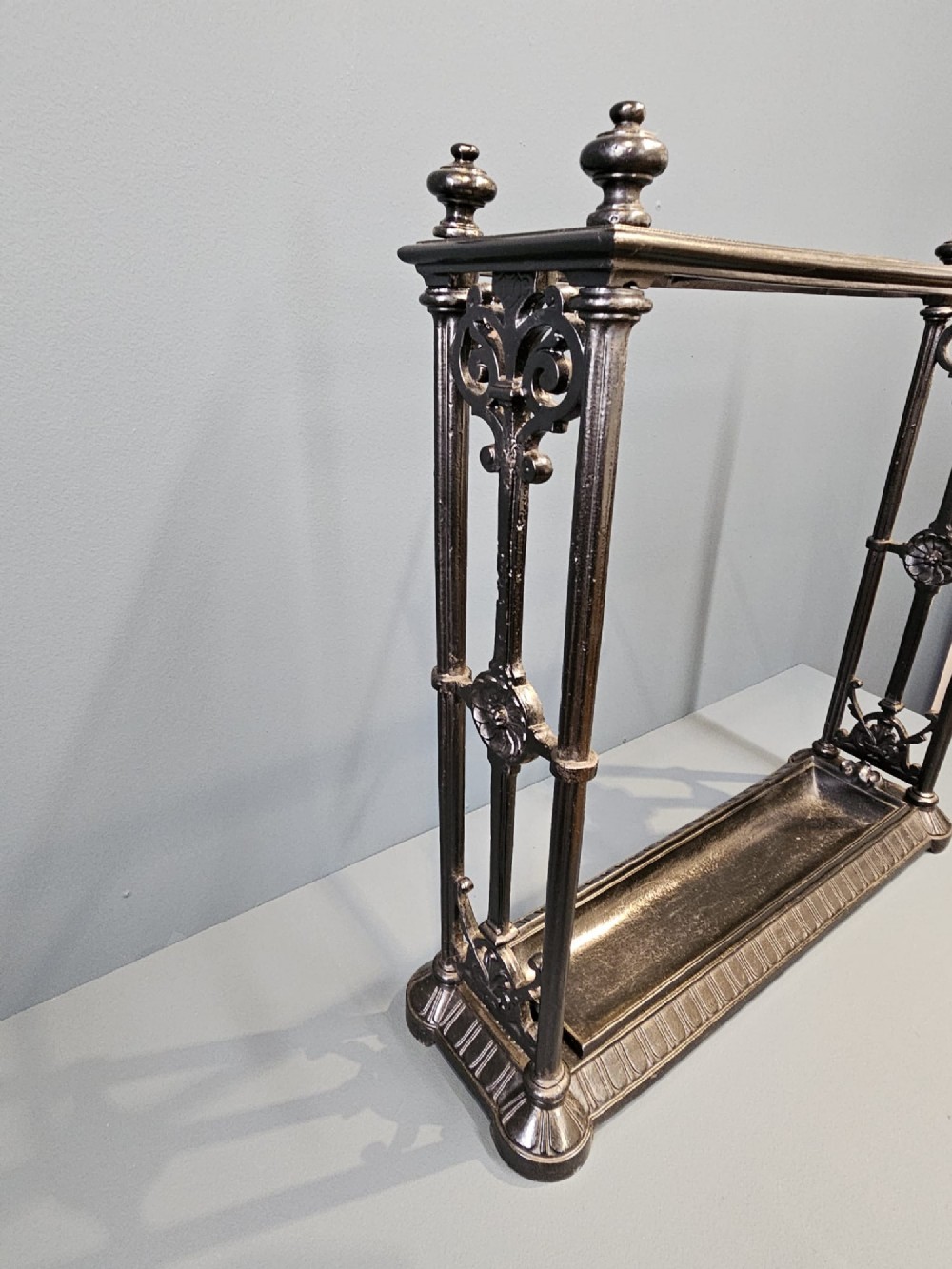 A rectangular Victorian cast iron stick stand featuring corner finials, multi-compartment top rail, vertical column supports, and a drip tray base. View VI