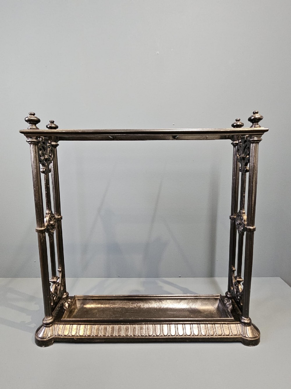 A rectangular Victorian cast iron stick stand featuring corner finials, multi-compartment top rail, vertical column supports, and a drip tray base. View VIII