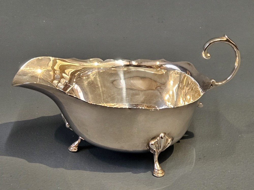 A George V polished sterling silver sauce boat featuring a wavy scalloped rim, a flying scroll handle, and three shell-capped feet.