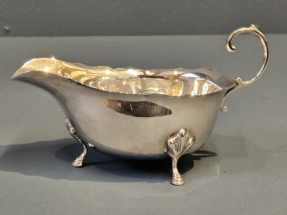 A George V polished sterling silver sauce boat featuring a wavy scalloped rim, a flying scroll handle, and three shell-capped feet. View II