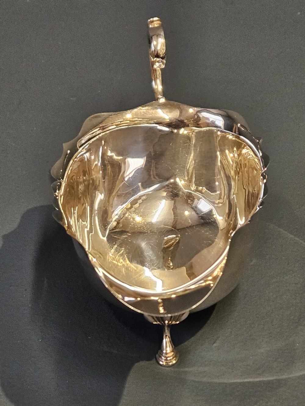 A George V polished sterling silver sauce boat featuring a wavy scalloped rim, a flying scroll handle, and three shell-capped feet. View IV