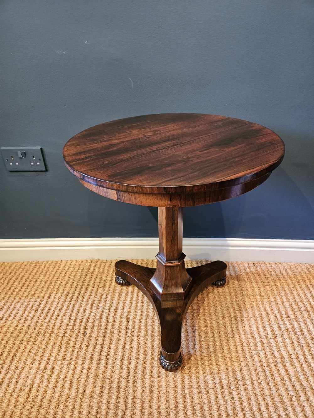 A round antique Regency rosewood occasional table featuring a rich grain, a central faceted pedestal column, and a tri-form platform base resting on carved feet.