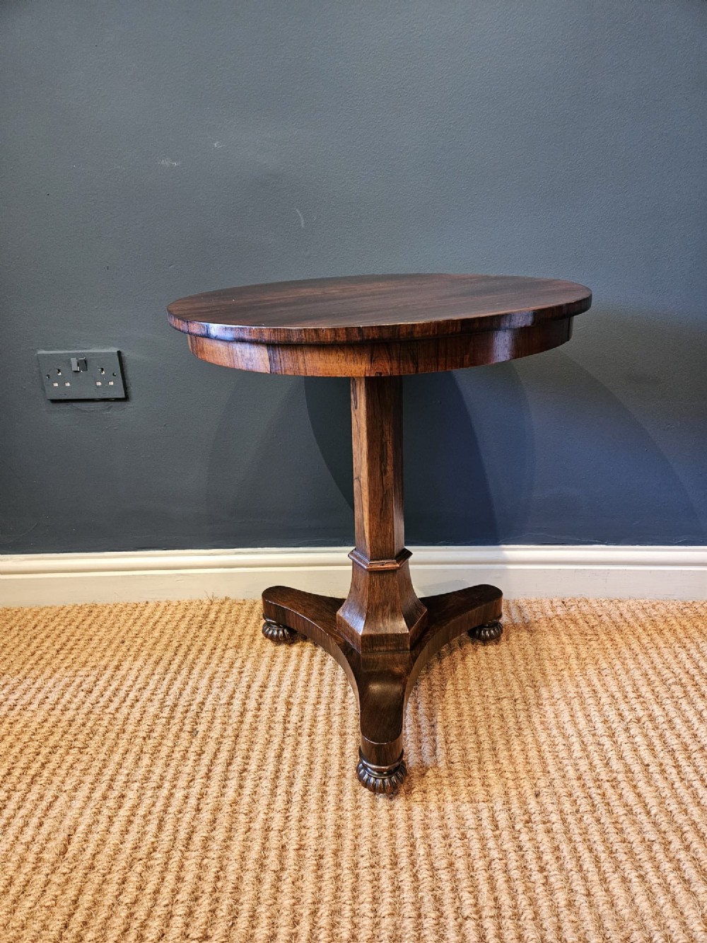 A round antique Regency rosewood occasional table featuring a rich grain, a central faceted pedestal column, and a tri-form platform base resting on carved feet. View II