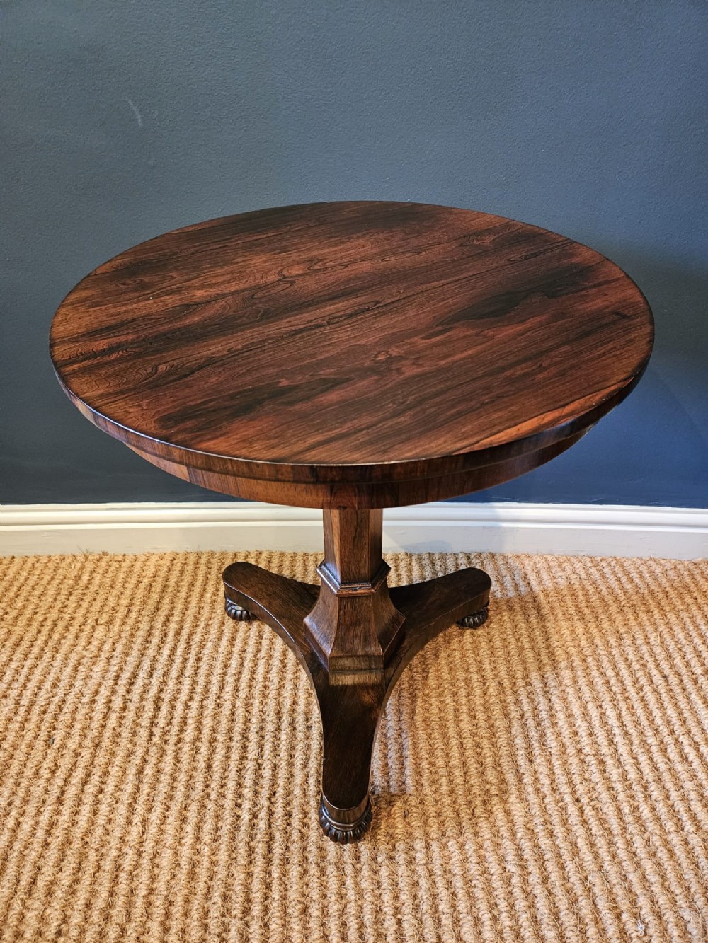 A round antique Regency rosewood occasional table featuring a rich grain, a central faceted pedestal column, and a tri-form platform base resting on carved feet. View V