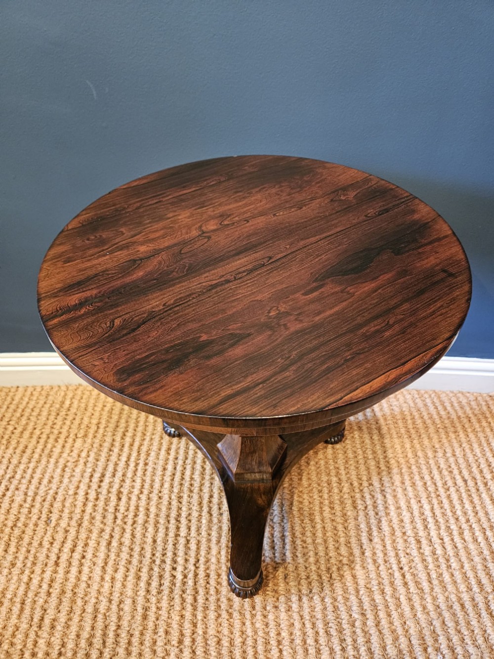 A round antique Regency rosewood occasional table featuring a rich grain, a central faceted pedestal column, and a tri-form platform base resting on carved feet. View VI