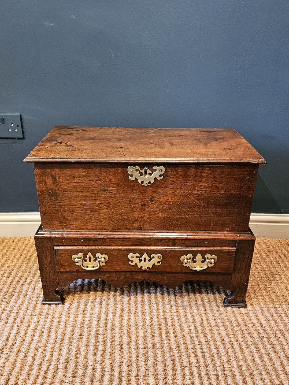 An antique 18th-century George II Welsh oak coffer bach with a hinged top lid, lower drawer, shaped apron, and brass escutcheons.