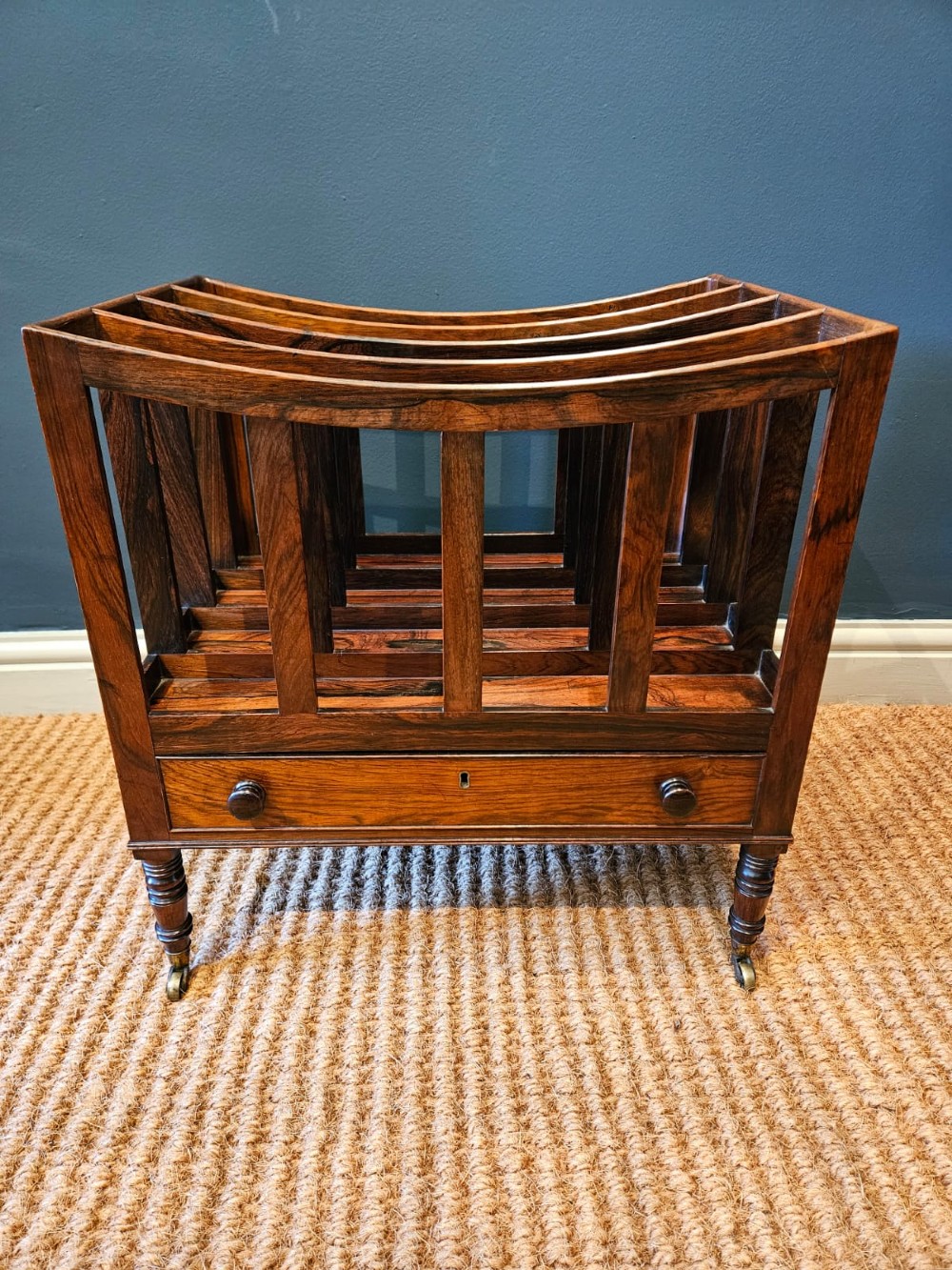 An antique Regency rosewood music canterbury featuring four slatted divisions with concave top rails, a single base drawer with turned wooden knobs, and turned legs on brass castors. View II