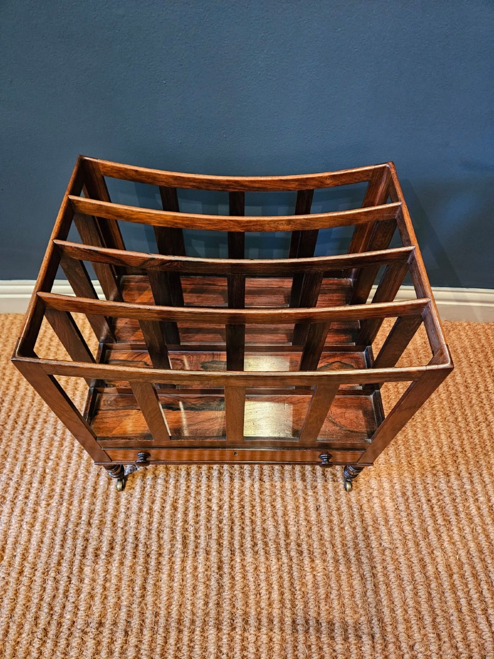 An antique Regency rosewood music canterbury featuring four slatted divisions with concave top rails, a single base drawer with turned wooden knobs, and turned legs on brass castors. View VI
