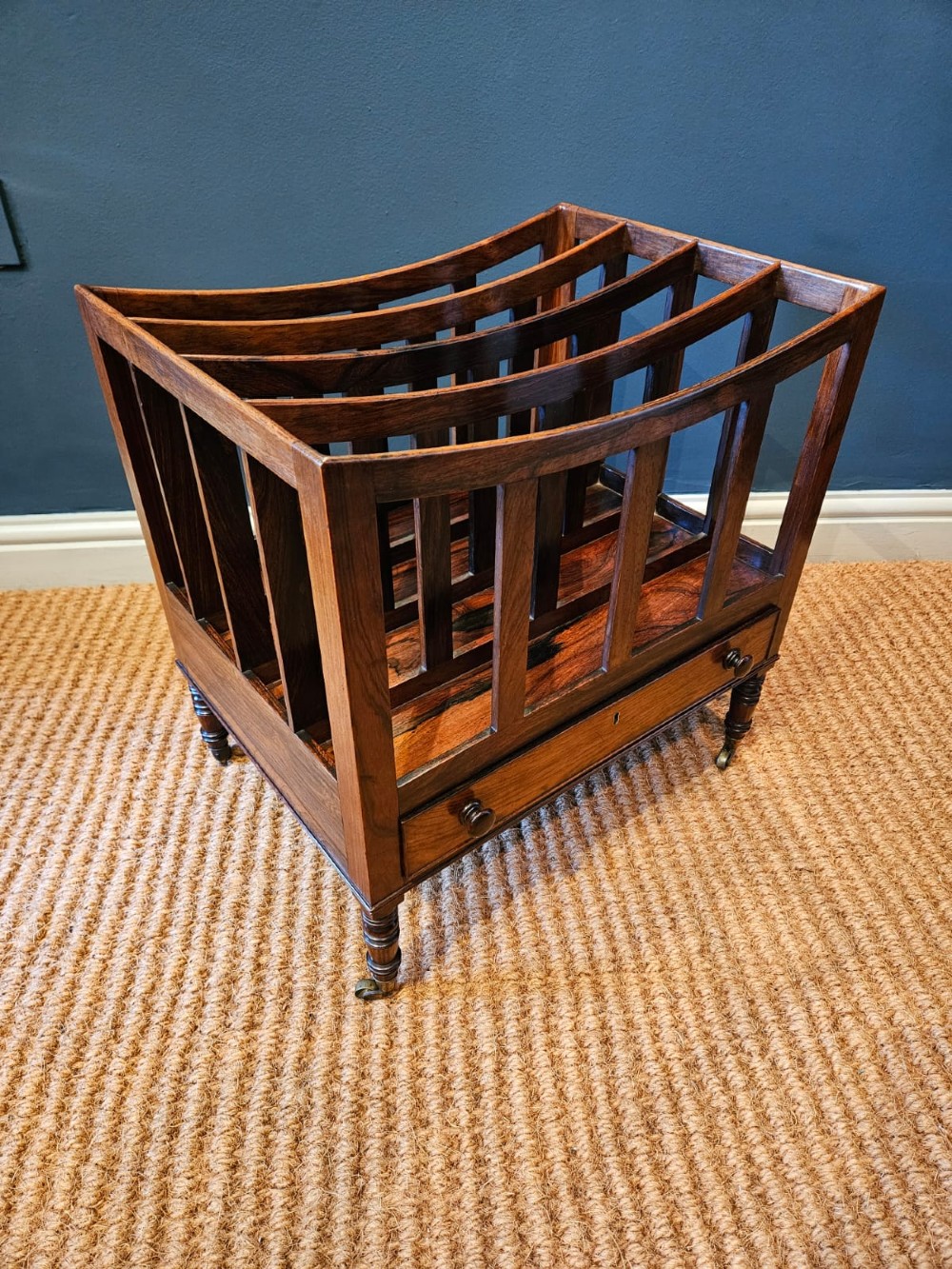 An antique Regency rosewood music canterbury featuring four slatted divisions with concave top rails, a single base drawer with turned wooden knobs, and turned legs on brass castors. View VII