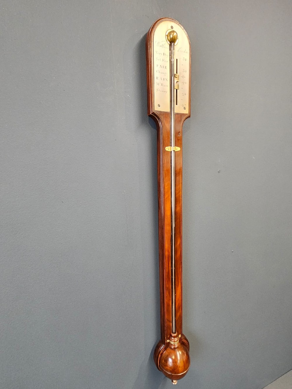 A wall-mounted Georgian mahogany stick barometer featuring a glass mercury tube, silvered brass register plate, and a bulbous cistern cover at the base.