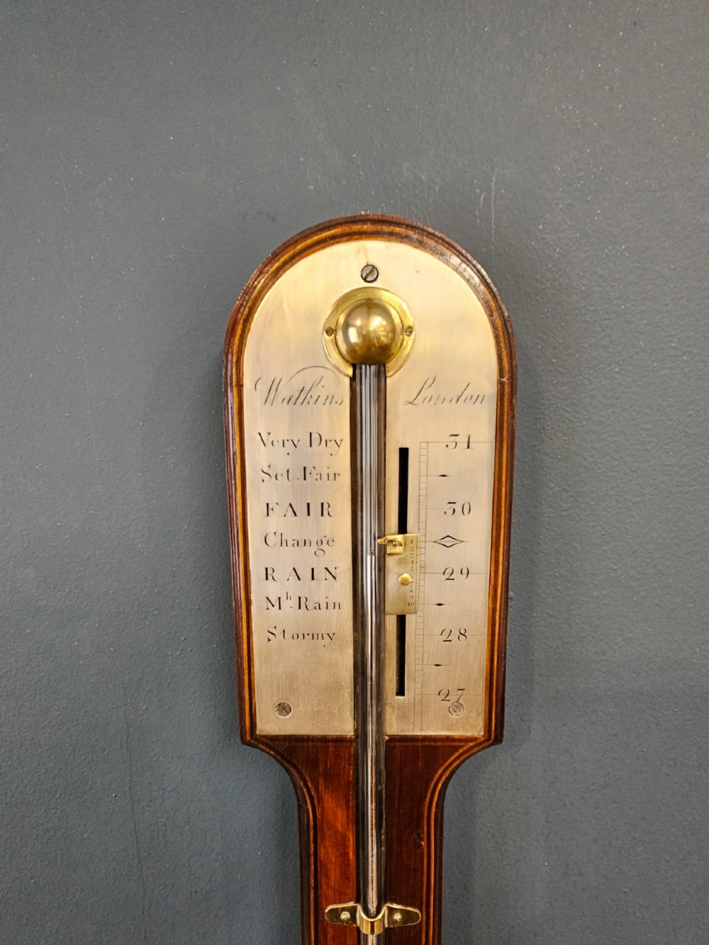 A wall-mounted Georgian mahogany stick barometer featuring a glass mercury tube, silvered brass register plate, and a bulbous cistern cover at the base. View V