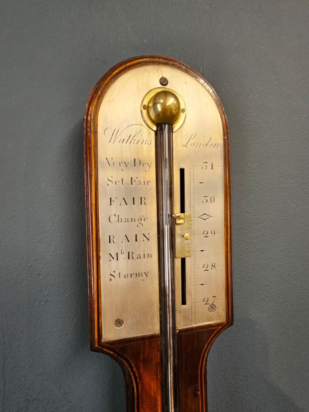 A wall-mounted Georgian mahogany stick barometer featuring a glass mercury tube, silvered brass register plate, and a bulbous cistern cover at the base. View VI