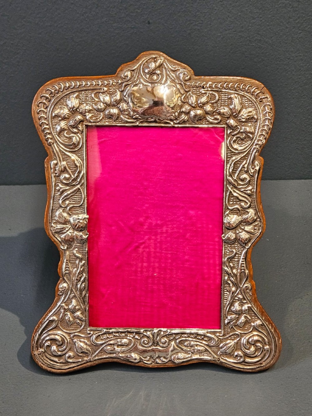An Edwardian Art Nouveau silver photo frame featuring embossed floral scrollwork mounted on a shaped wooden backing with a bright pink interior insert.