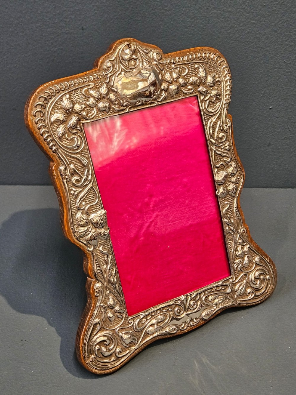 An Edwardian Art Nouveau silver photo frame featuring embossed floral scrollwork mounted on a shaped wooden backing with a bright pink interior insert. View II