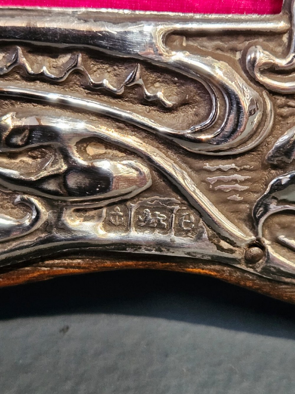 An Edwardian Art Nouveau silver photo frame featuring embossed floral scrollwork mounted on a shaped wooden backing with a bright pink interior insert. View V