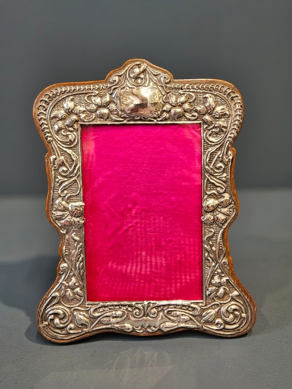 An Edwardian Art Nouveau silver photo frame featuring embossed floral scrollwork mounted on a shaped wooden backing with a bright pink interior insert. View VI