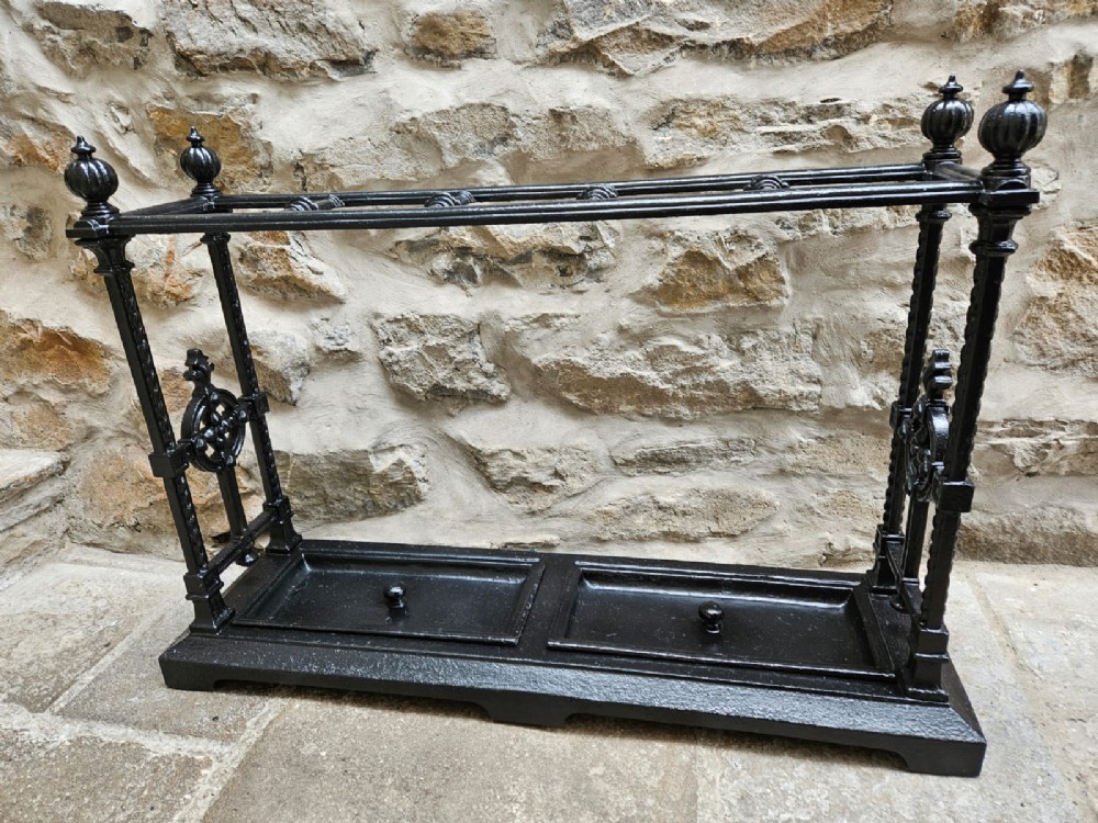 A black cast iron Victorian stick stand featuring finial top ornamentation, side supports, and two removable drip trays at the base.