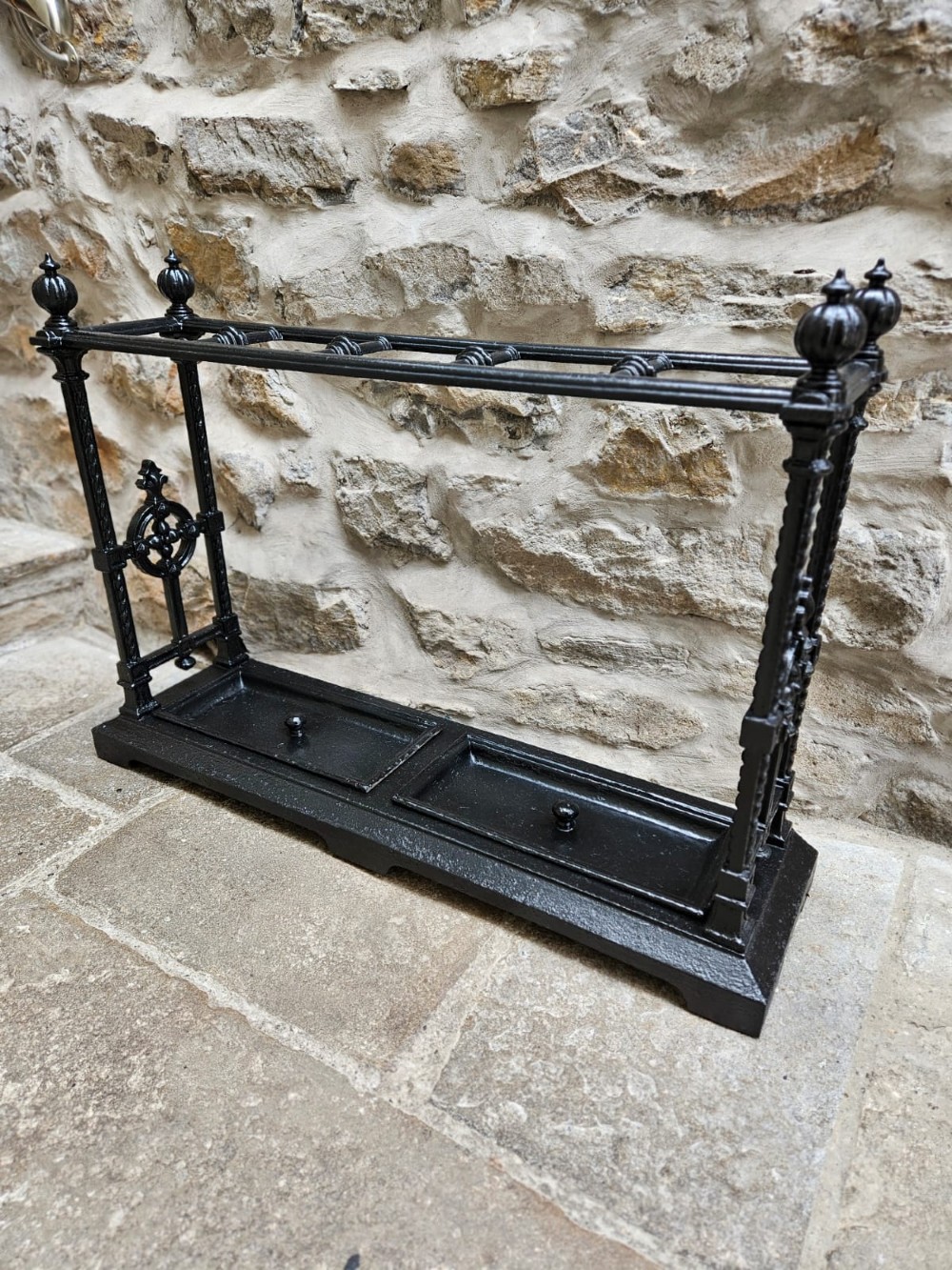 A black cast iron Victorian stick stand featuring finial top ornamentation, side supports, and two removable drip trays at the base. View III
