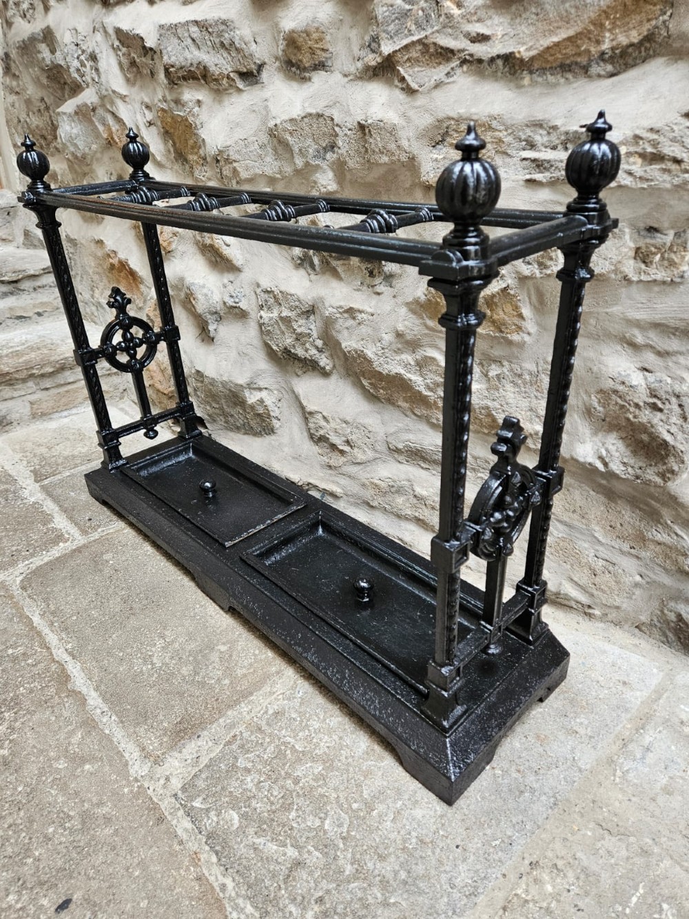 A black cast iron Victorian stick stand featuring finial top ornamentation, side supports, and two removable drip trays at the base. View IV