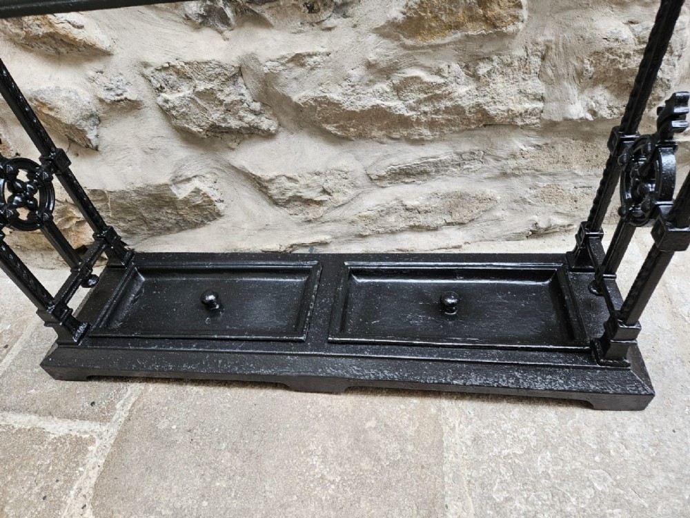 A black cast iron Victorian stick stand featuring finial top ornamentation, side supports, and two removable drip trays at the base. View V