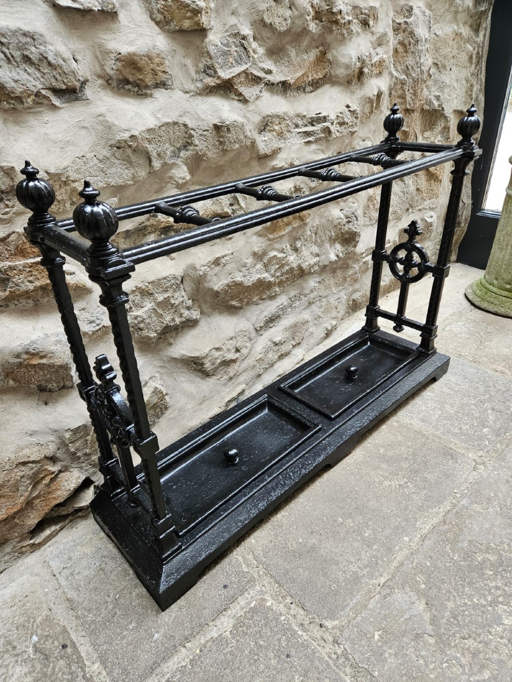 A black cast iron Victorian stick stand featuring finial top ornamentation, side supports, and two removable drip trays at the base. View VI