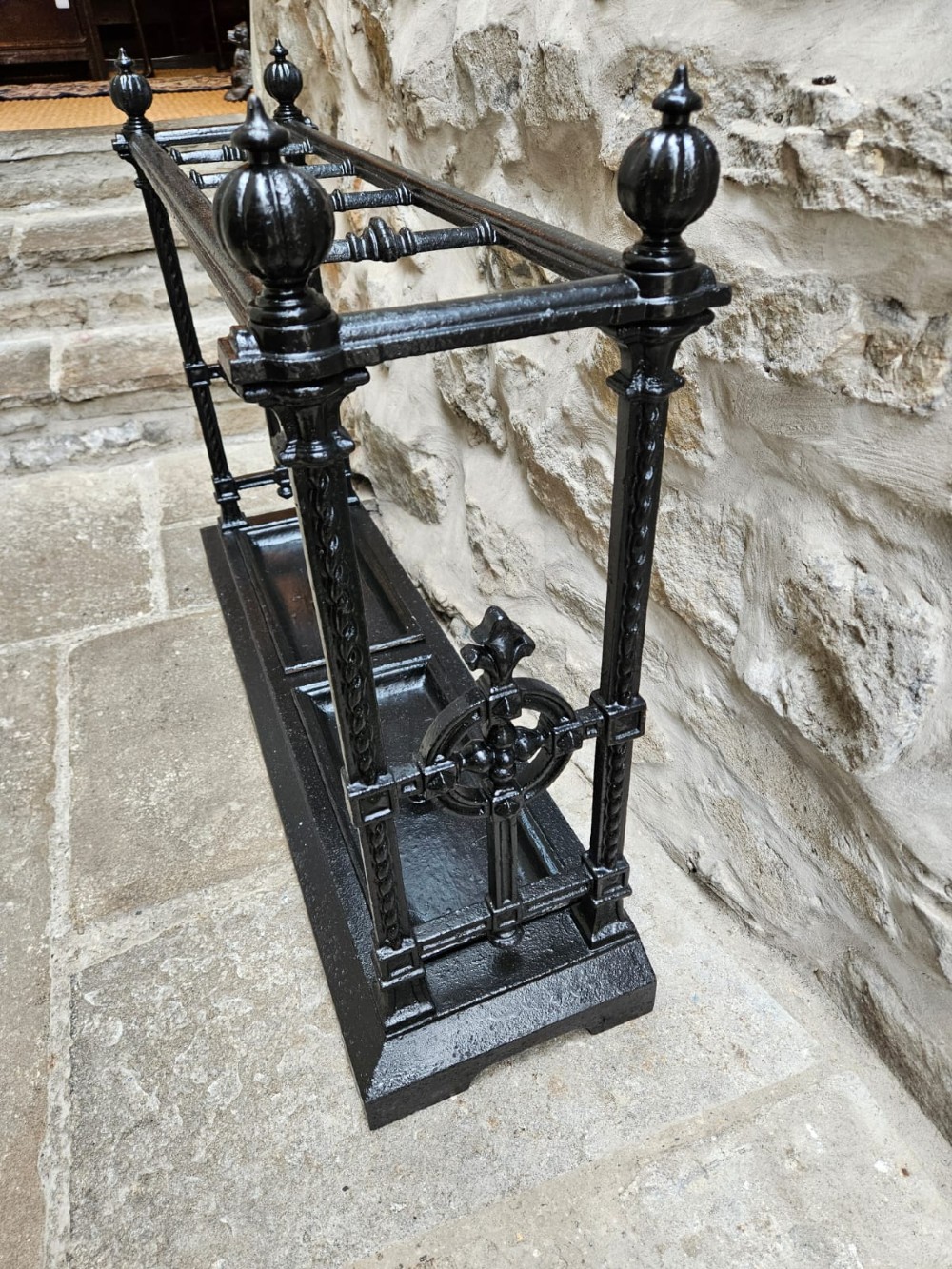 A black cast iron Victorian stick stand featuring finial top ornamentation, side supports, and two removable drip trays at the base. View VII