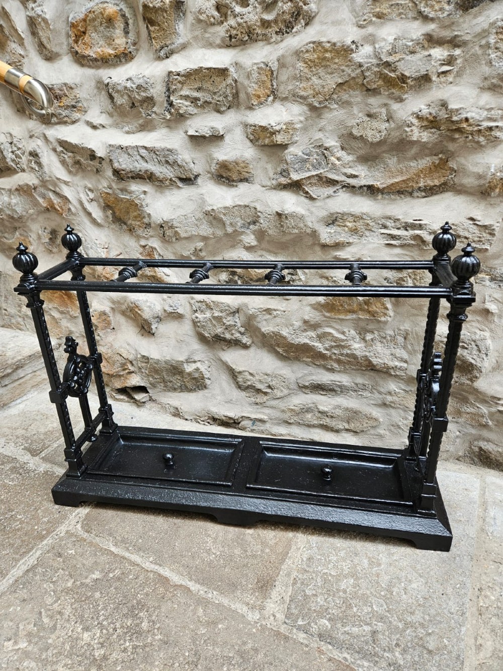 A black cast iron Victorian stick stand featuring finial top ornamentation, side supports, and two removable drip trays at the base. View IX