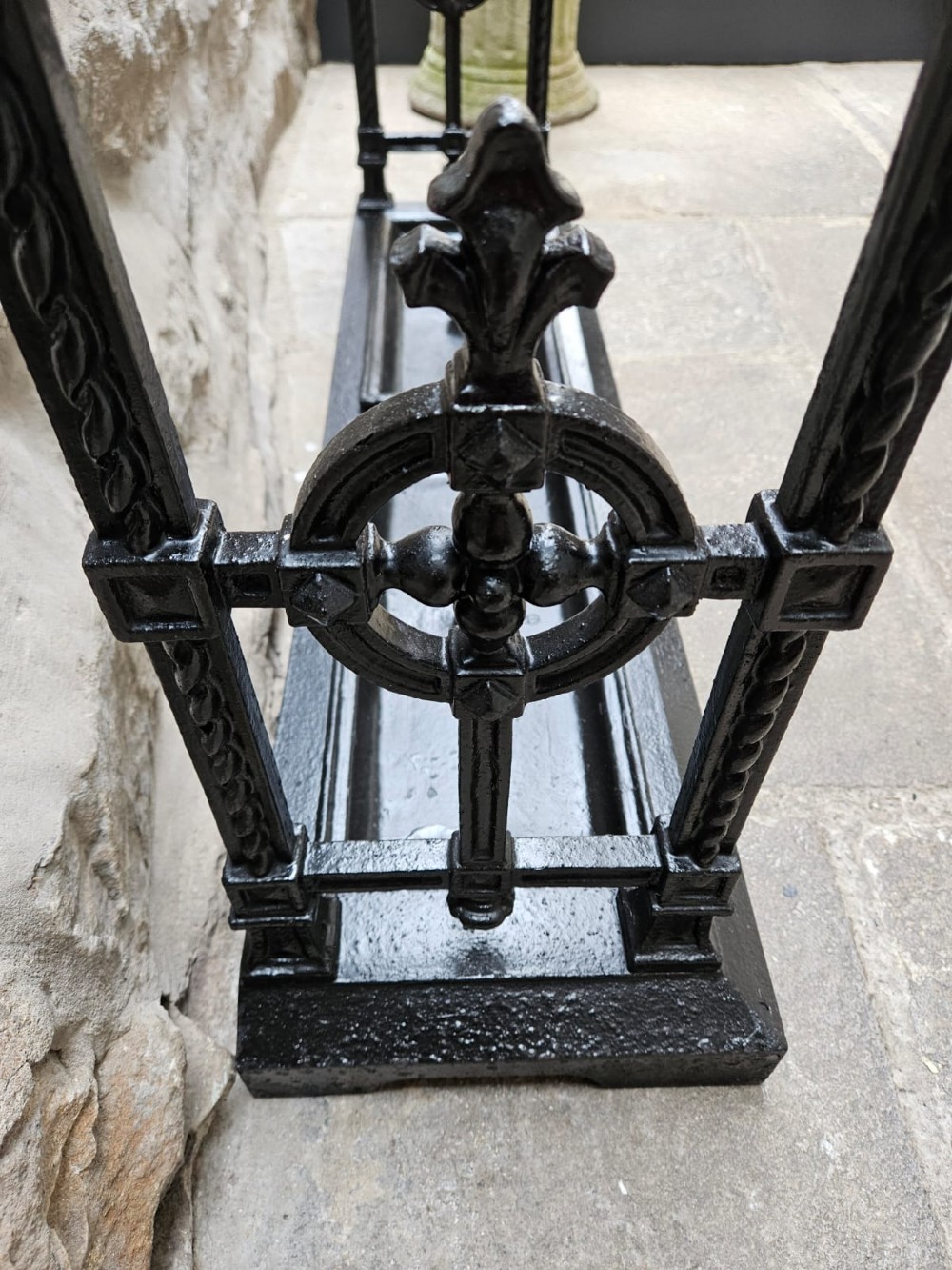 A black cast iron Victorian stick stand featuring finial top ornamentation, side supports, and two removable drip trays at the base. View X