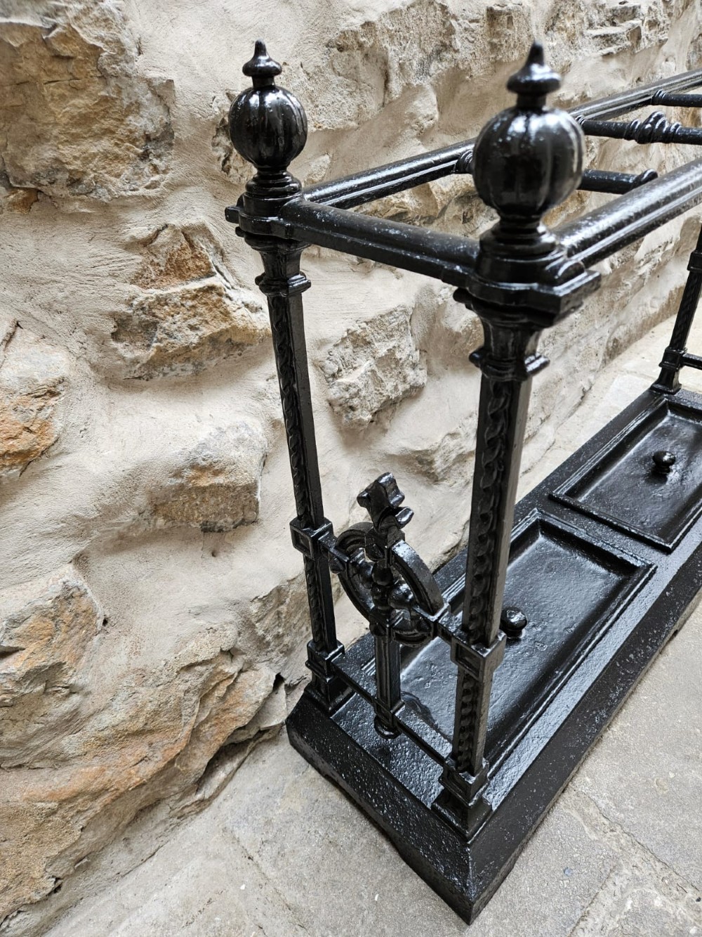 A black cast iron Victorian stick stand featuring finial top ornamentation, side supports, and two removable drip trays at the base. View XI