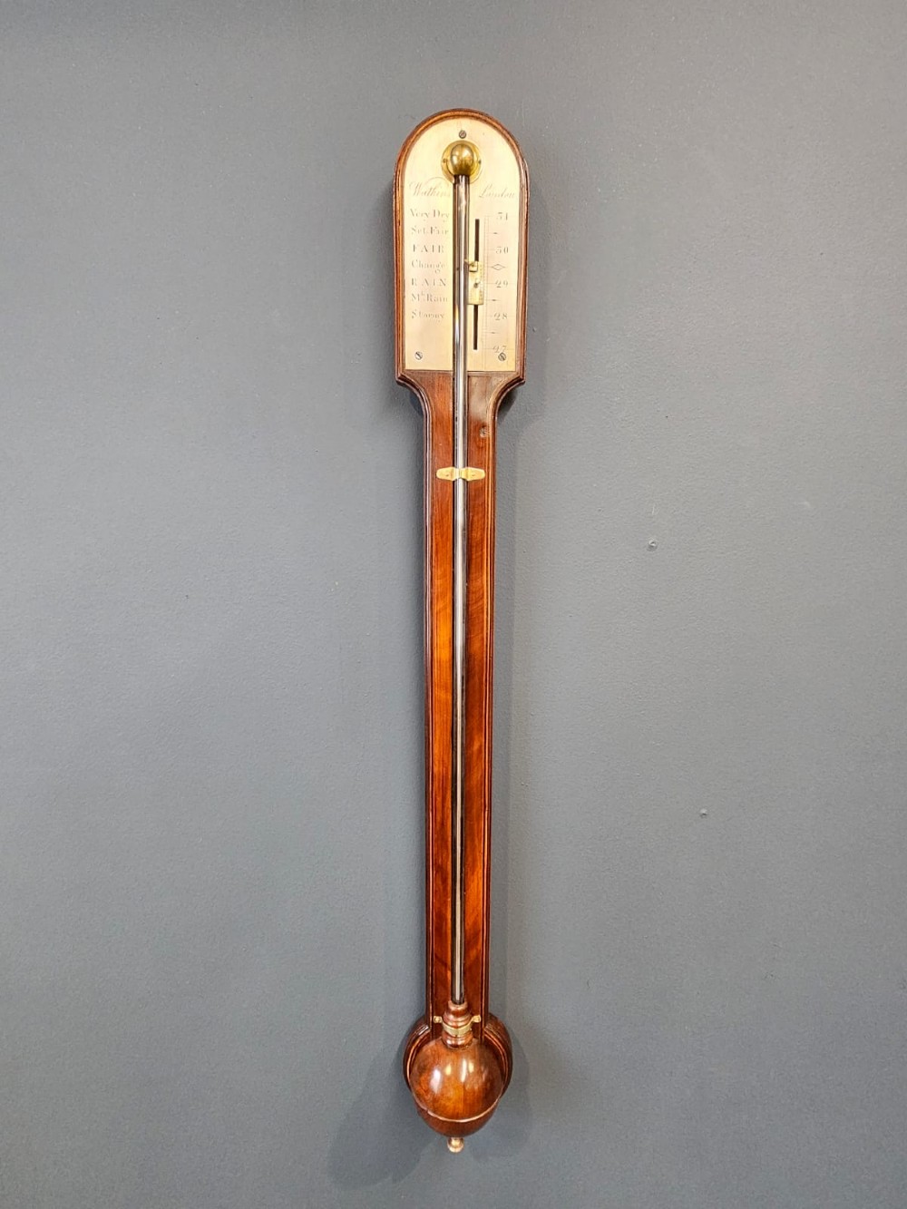 A wall-mounted Georgian mahogany stick barometer featuring a capillary glass tube, engraved silvered register plate, and a rounded wooden cistern cover at the base. View III