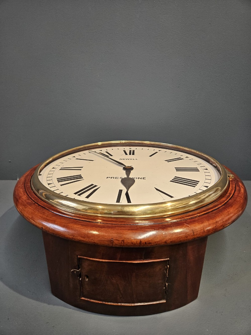 A circular Victorian wall clock featuring a mahogany wooden frame, a white dial with black Roman numerals, spade hands, and a brass bezel. View V